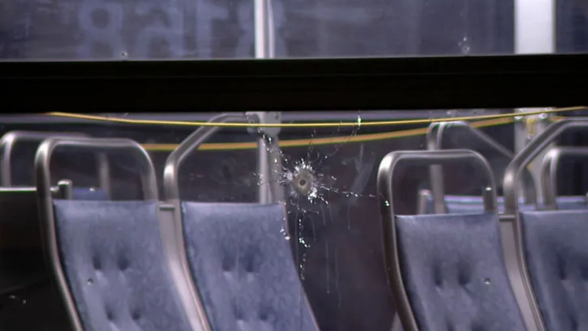 Bullet hole in a window with glass shatter and spray marks inside a vehicle with blue upholstered seats and metal frames.