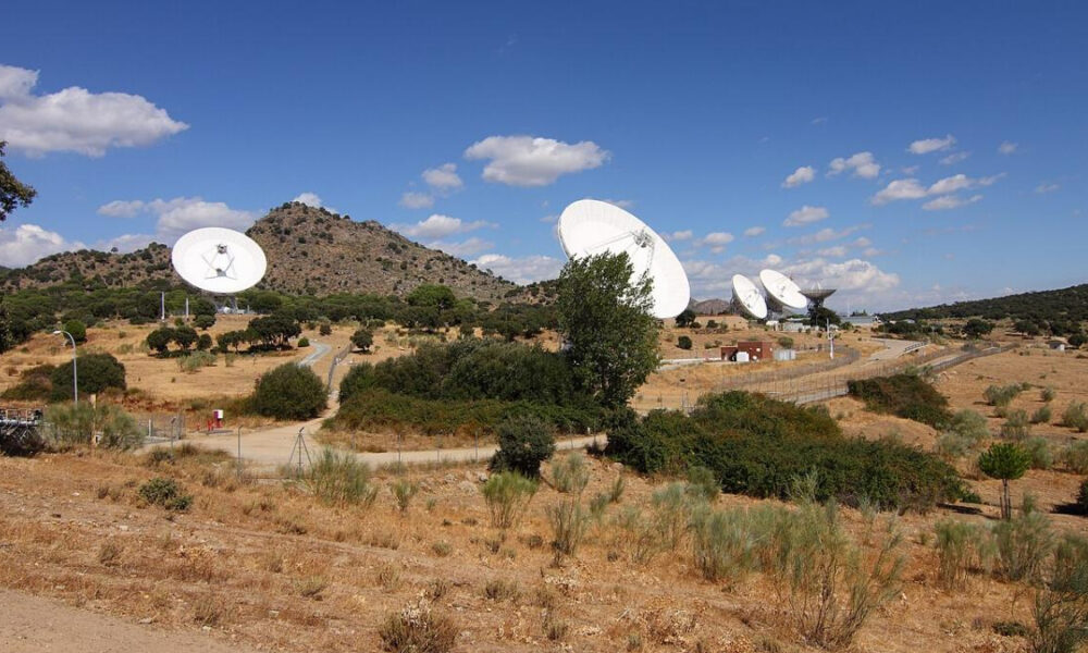 A desert landscape with several large white satellite dishes facing outward and a blue sky above hills in the distance.