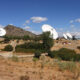 A desert landscape with several large white satellite dishes facing outward and a blue sky above hills in the distance.
