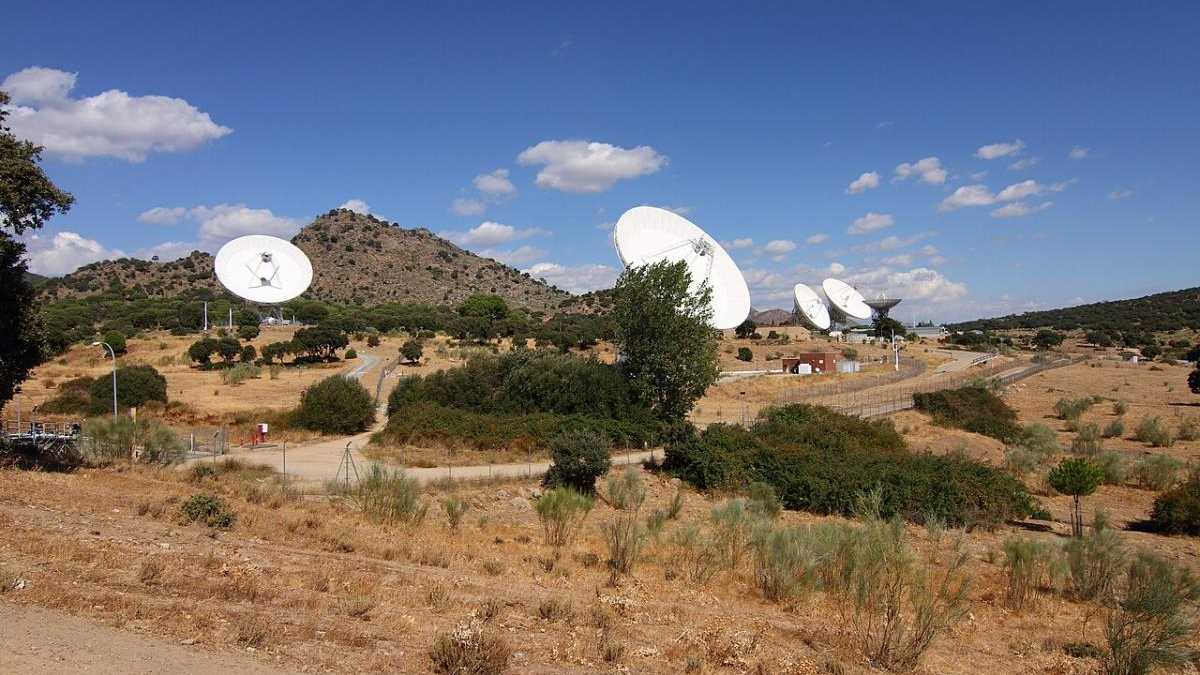 A desert landscape with several large white satellite dishes facing outward and a blue sky above hills in the distance.
