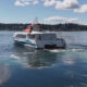 Passenger ferry boat sailing on calm blue water with shoreline and trees in the background under a clear sky.