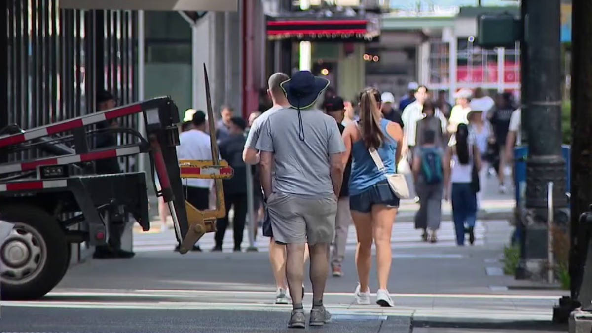 Man wearing a wide-brimmed hat and gray shirt walks with a woman in a blue tank top across a busy city crosswalk; construction truck visible on the left.