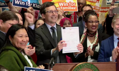 Smiling man in a suit holds up a signed document at a press conference, surrounded by supporters with 'Win for Kids' signs in the background.