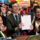 Smiling man in a suit holds up a signed document at a press conference, surrounded by supporters with 'Win for Kids' signs in the background.