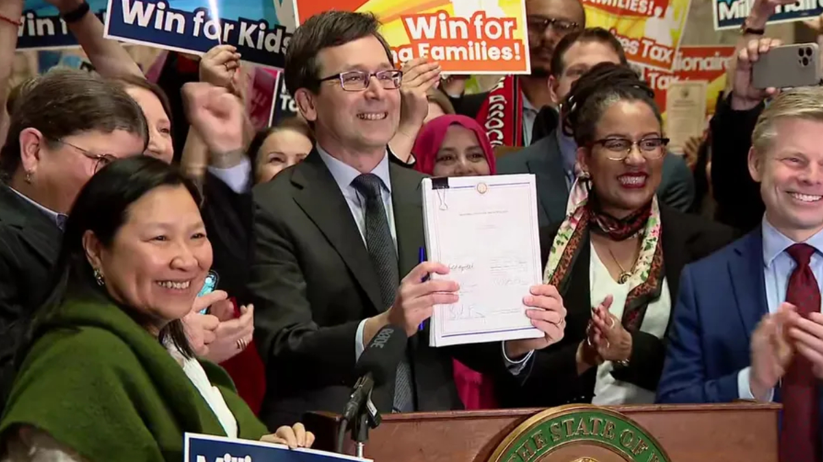 Smiling man in a suit holds up a signed document at a press conference, surrounded by supporters with 'Win for Kids' signs in the background.