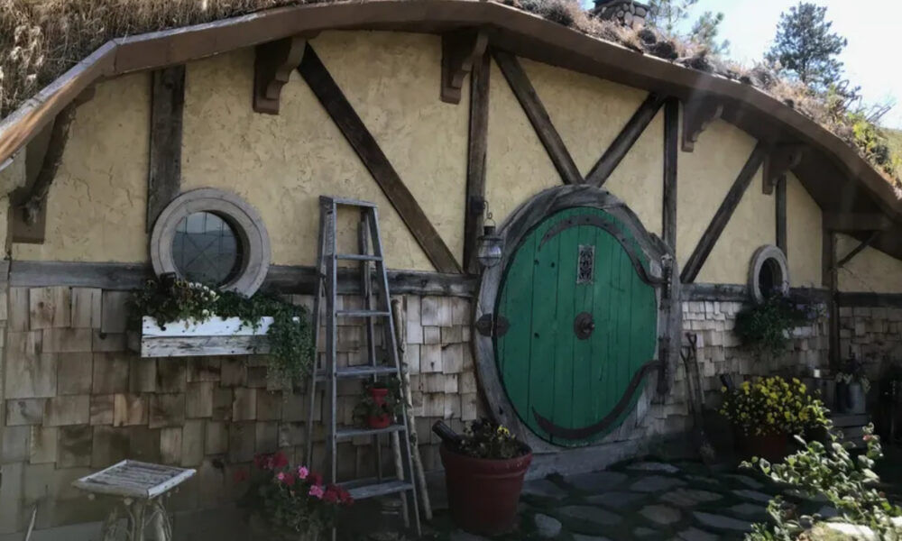 Hobbit-style cottage wall with a large round green door, timber beams, and potted plants.