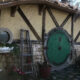 Hobbit-style cottage wall with a large round green door, timber beams, and potted plants.