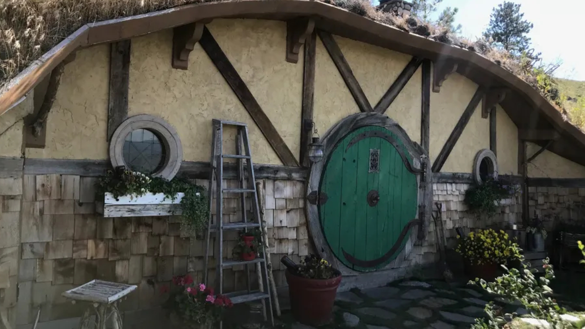 Hobbit-style cottage wall with a large round green door, timber beams, and potted plants.