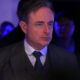 Middle-aged man in a gray suit and polka-dot tie at a dim, blue-lit event, looking to the right.