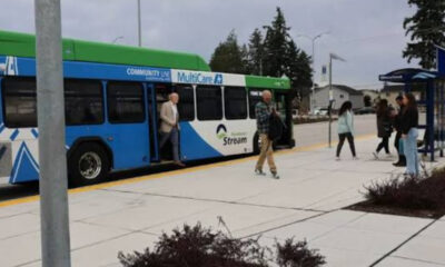 Blue and green community bus with open doors at a curb, several people boarding and waiting nearby at a bus stop.
