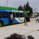 Blue and green community bus with open doors at a curb, several people boarding and waiting nearby at a bus stop.