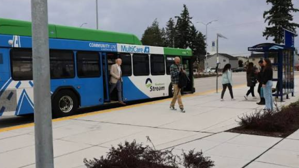 Blue and green community bus with open doors at a curb, several people boarding and waiting nearby at a bus stop.