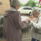 Two elderly women stand on a sidewalk by a railing, one aiding the other as they hold hands near a parked car and houses in a suburban street.