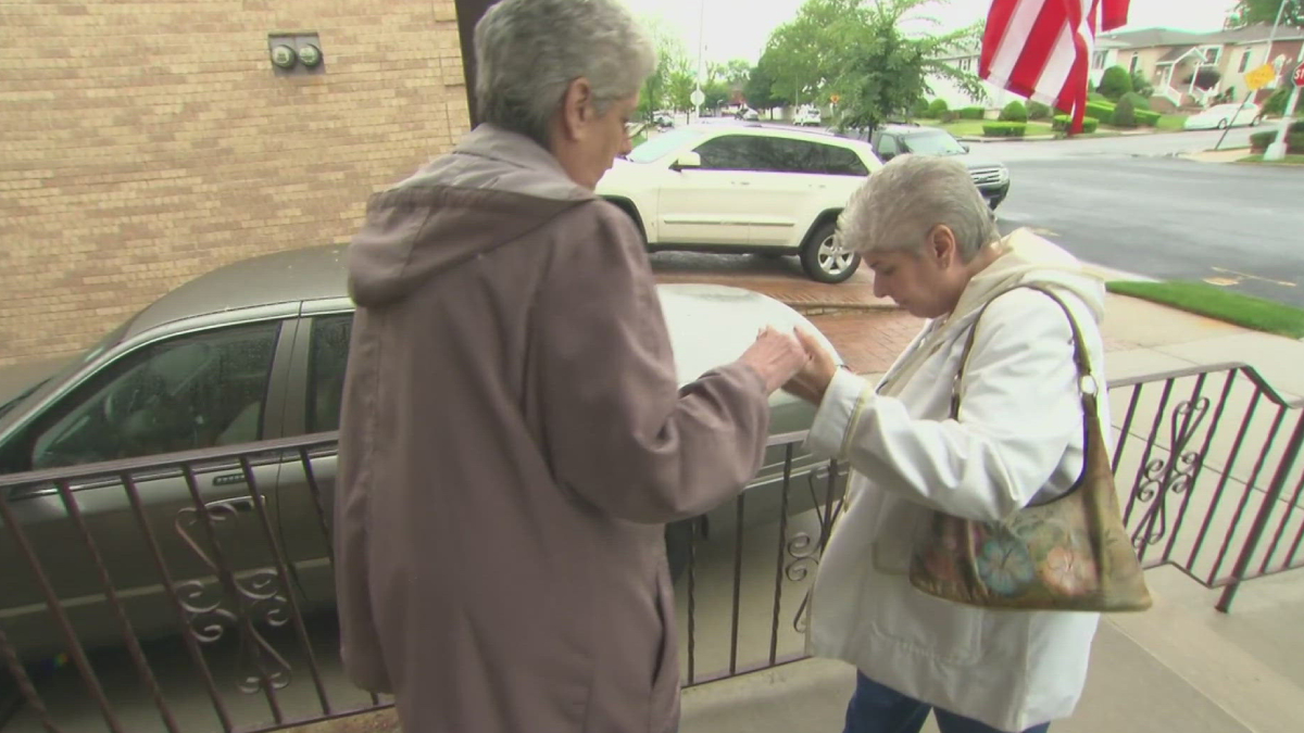 Two elderly women stand on a sidewalk by a railing, one aiding the other as they hold hands near a parked car and houses in a suburban street.
