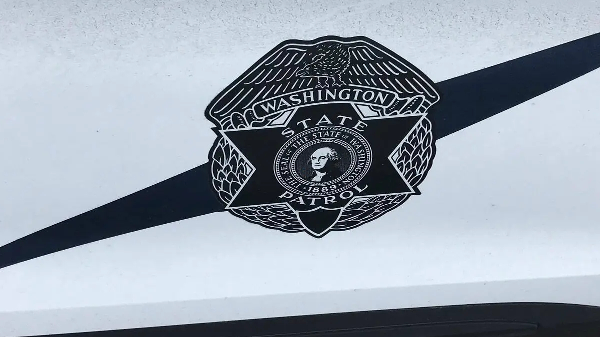 Close-up of a Washington State Patrol badge emblem on a white vehicle with a bold black diagonal stripe behind it.