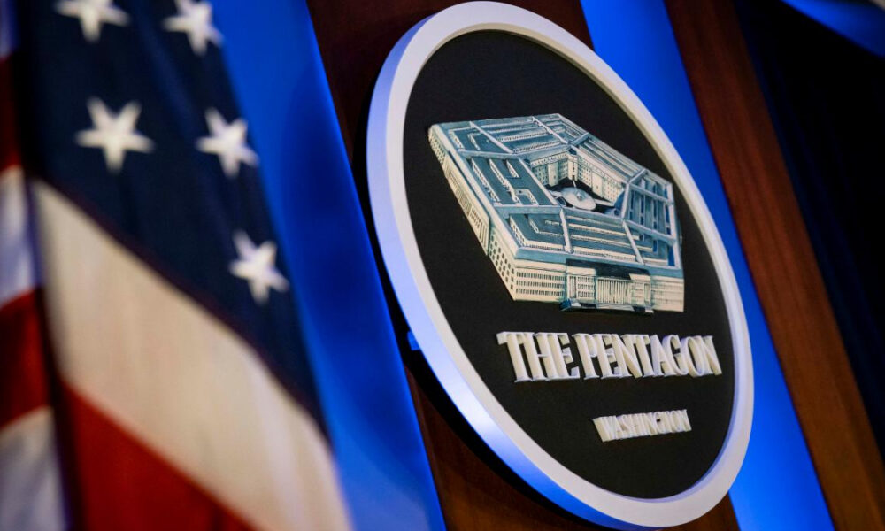 Circular The Pentagon emblem on a podium, with an American flag in the background under blue lighting.