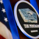 Circular The Pentagon emblem on a podium, with an American flag in the background under blue lighting.