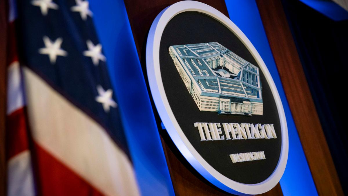 Circular The Pentagon emblem on a podium, with an American flag in the background under blue lighting.