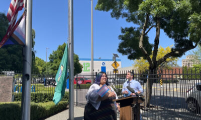 Two presenters stand at a wooden lectern outdoors beside a black fence, with flags and a green banner nearby on a sunny day.
