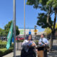 Two presenters stand at a wooden lectern outdoors beside a black fence, with flags and a green banner nearby on a sunny day.