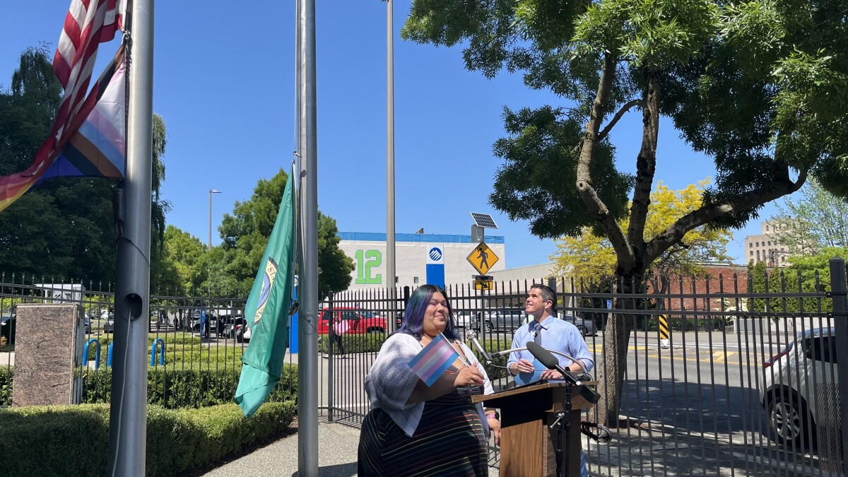 Two presenters stand at a wooden lectern outdoors beside a black fence, with flags and a green banner nearby on a sunny day.