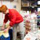Volunteer in a red sweatshirt bends over to open a bag of pet food in a cluttered donation room with shelves of supplies nearby.