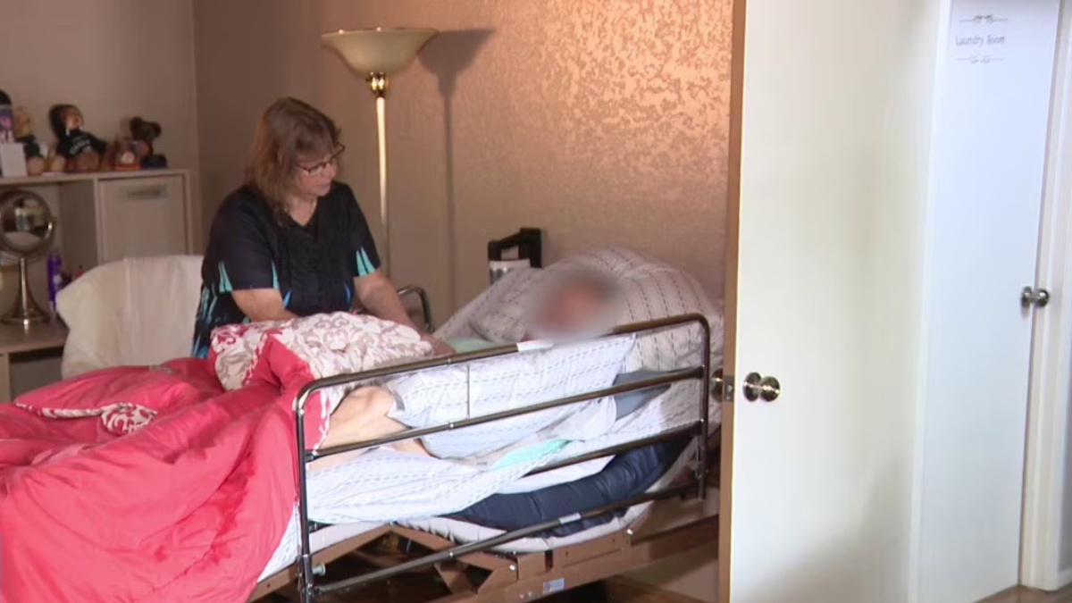 Caregiver sits beside a patient lying in a metal-frame bed with pink and white bedding in a cozy bedroom.