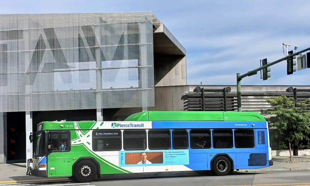 Public transit bus (Pierce Transit) green and blue, driving past a modern, mesh-covered building on a city street.