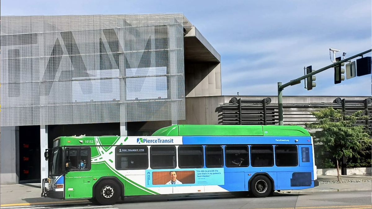 Public transit bus (Pierce Transit) green and blue, driving past a modern, mesh-covered building on a city street.