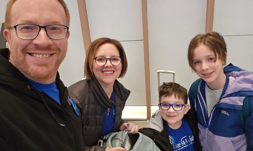 A family of four posing for a selfie in an airport terminal before their flight, smiling at the camera.