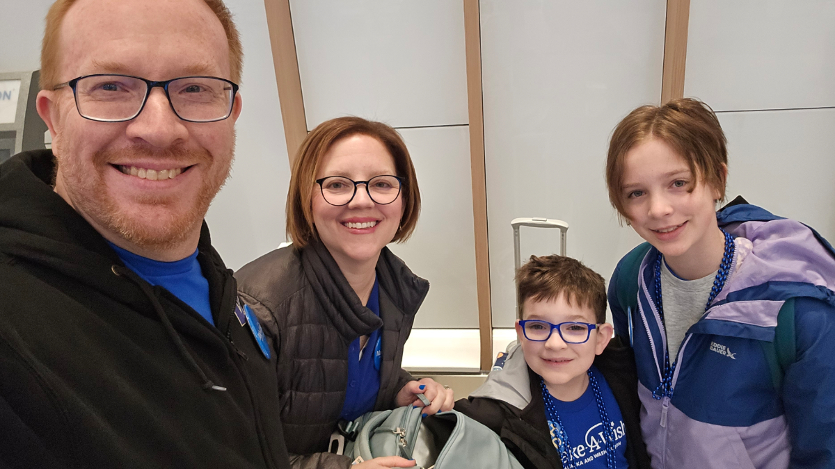 A family of four posing for a selfie in an airport terminal before their flight, smiling at the camera.