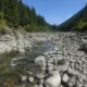 Rugged riverbed with large gray rocks and clear flowing water flanked by forested slopes under a blue sky