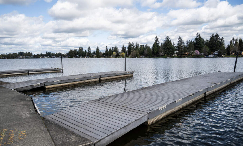 Floating wooden docks extend into a calm lake with a tree-lined shore and houses in the distance, under a cloudy sky.