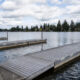 Floating wooden docks extend into a calm lake with a tree-lined shore and houses in the distance, under a cloudy sky.