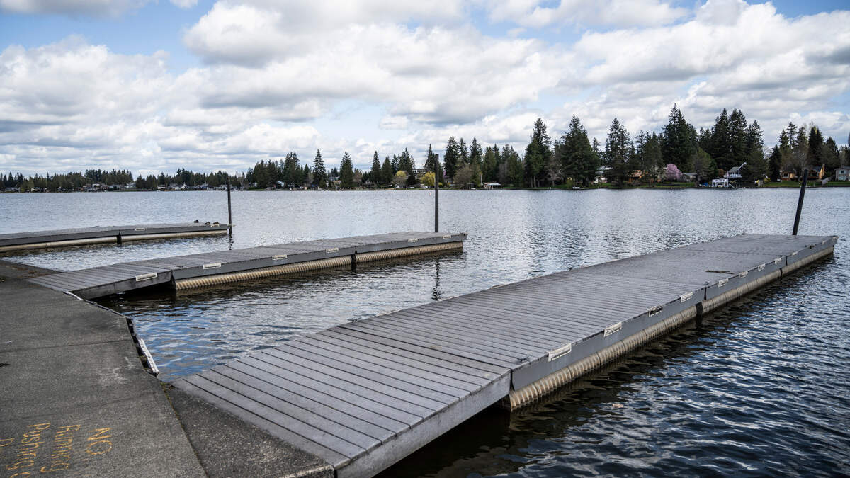 Floating wooden docks extend into a calm lake with a tree-lined shore and houses in the distance, under a cloudy sky.