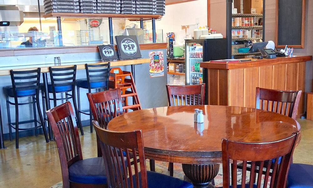 A cozy pizza shop interior with a round wooden table and six wooden chairs near a bar counter.