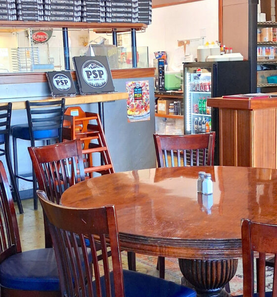 A cozy pizza shop interior with a round wooden table and six wooden chairs near a bar counter.