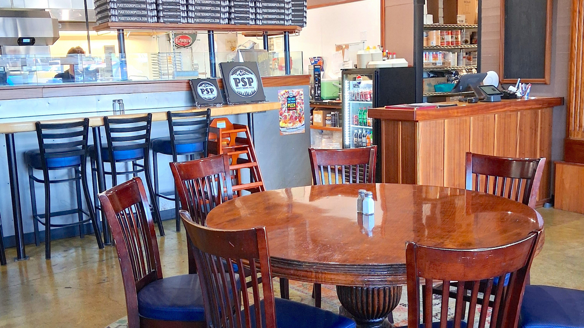 A cozy pizza shop interior with a round wooden table and six wooden chairs near a bar counter.