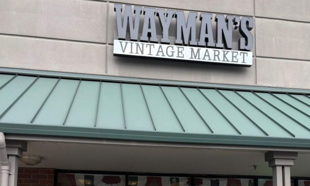 Storefront sign reading 'Wayman's Vintage Market' above a green metal awning on a gray brick façade, storefront visible below the awning.