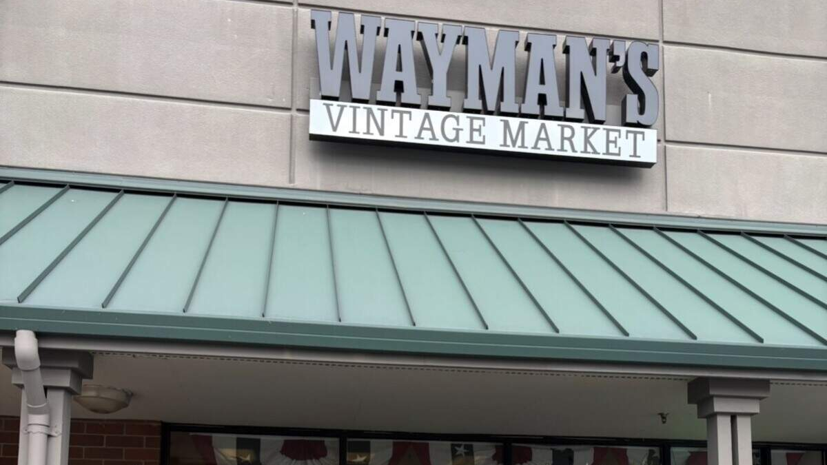 Storefront sign reading 'Wayman's Vintage Market' above a green metal awning on a gray brick façade, storefront visible below the awning.