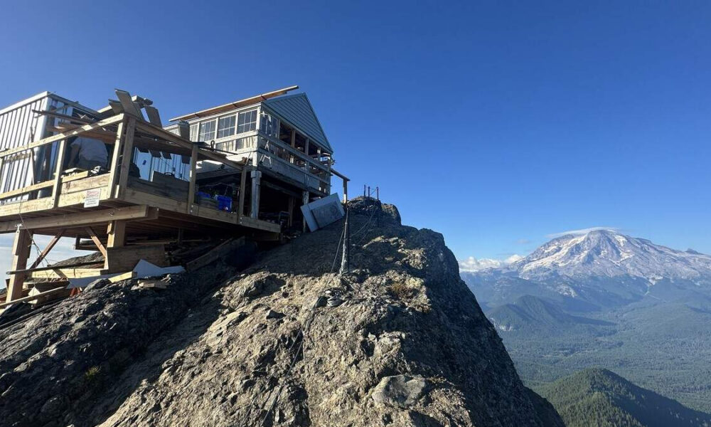 Wooden mountain hut with a railing on a rocky cliff, overlooking a broad valley and snow-capped mountains under a blue sky.