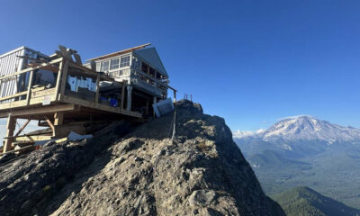 Wooden mountain hut with a railing on a rocky cliff, overlooking a broad valley and snow-capped mountains under a blue sky.
