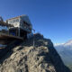 Wooden mountain hut with a railing on a rocky cliff, overlooking a broad valley and snow-capped mountains under a blue sky.