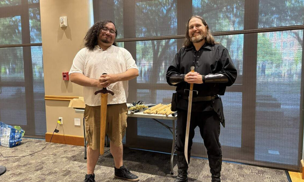 Two men stand indoors, each holding a sword upright in front of them; the man on the left wears a white polo and khaki shorts, the man on the right wears a black medieval costume with leather bracers and boots behind a table of props.