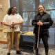 Two men stand indoors, each holding a sword upright in front of them; the man on the left wears a white polo and khaki shorts, the man on the right wears a black medieval costume with leather bracers and boots behind a table of props.