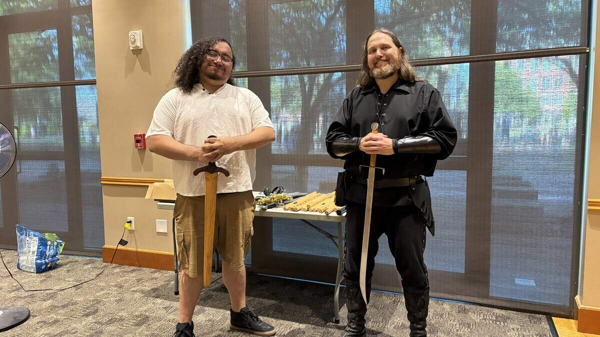 Two men stand indoors, each holding a sword upright in front of them; the man on the left wears a white polo and khaki shorts, the man on the right wears a black medieval costume with leather bracers and boots behind a table of props.