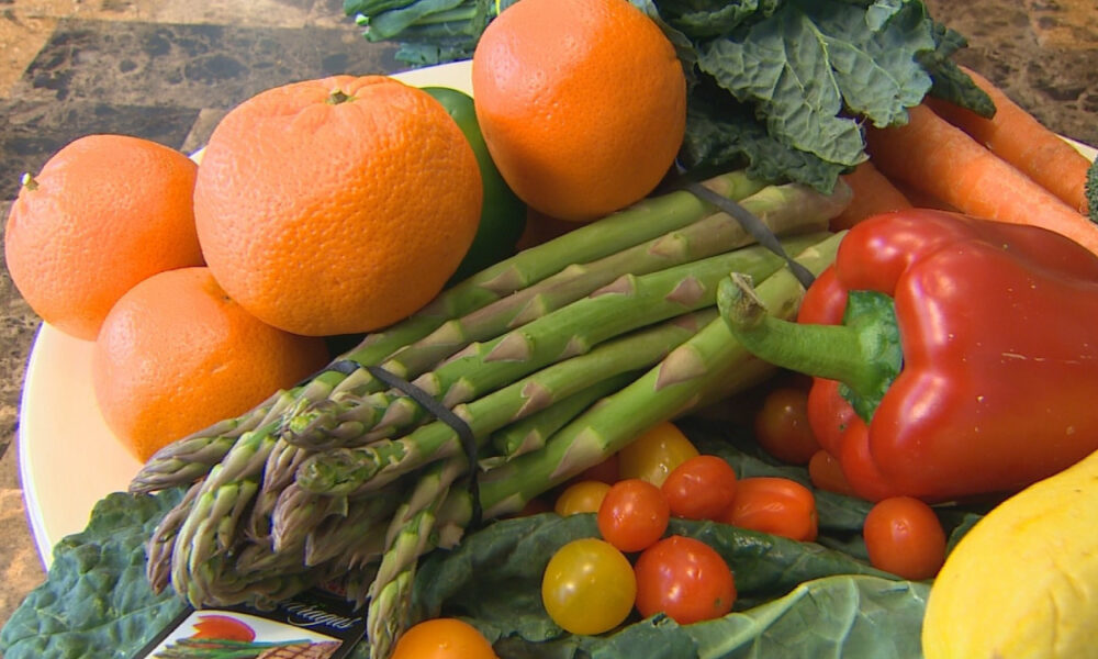 Assorted fresh vegetables and fruits: oranges, asparagus, a red bell pepper, cherry tomatoes, leafy greens, carrots, and broccoli on a plate.