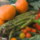 Assorted fresh vegetables and fruits: oranges, asparagus, a red bell pepper, cherry tomatoes, leafy greens, carrots, and broccoli on a plate.