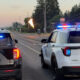 Two police SUVs with flashing blue and red lights parked on a road at dusk, flames rising in the distance as an orange torch or flare is visible on the street.
