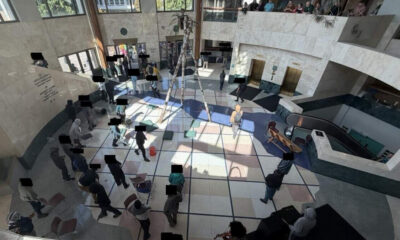 Overhead view of a spacious atrium with a central palm tree, sunlit tiled floor, and people lined up or gathered along the walls and seating areas.